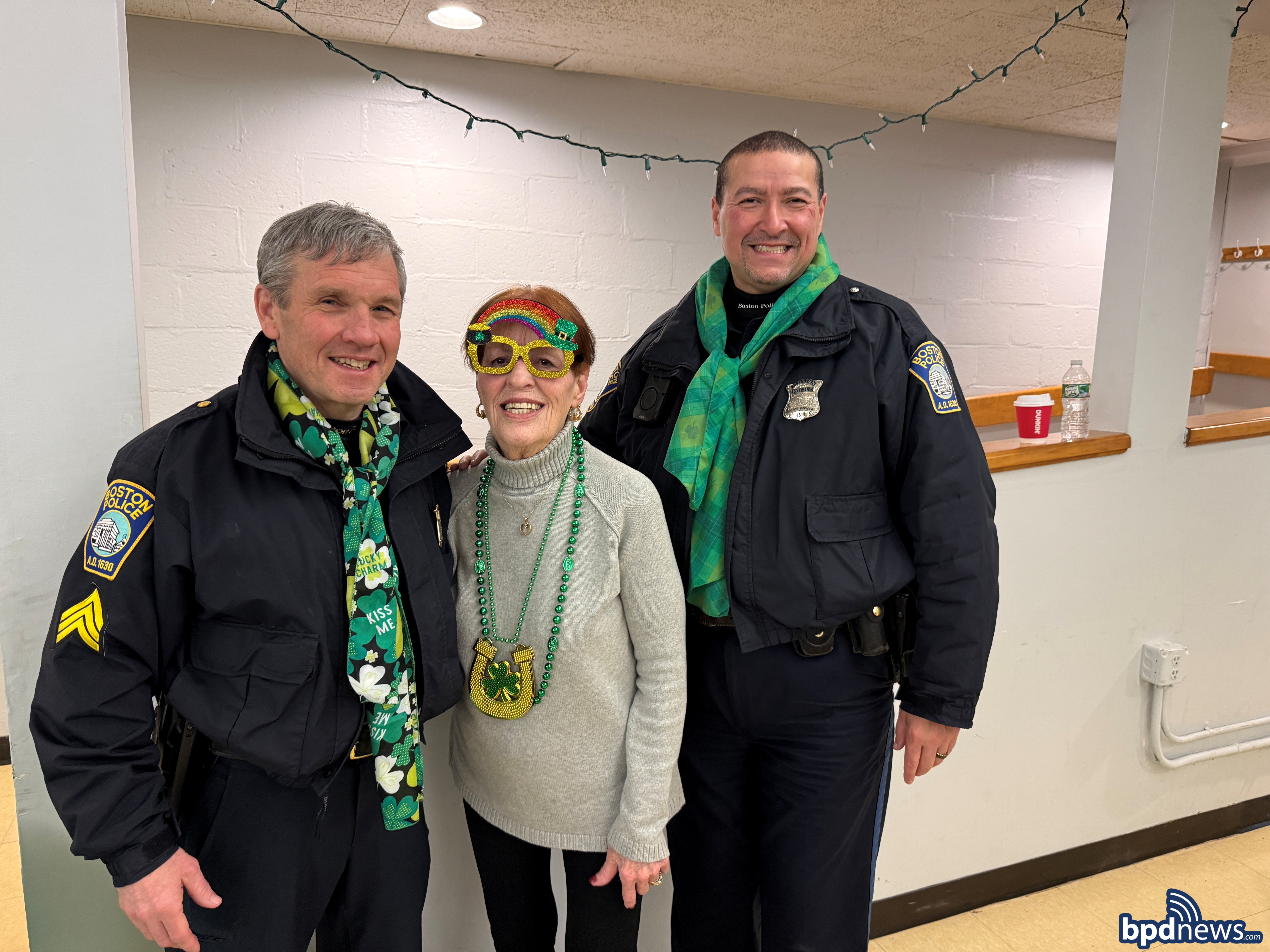 Two Boston Police Officers pose for a picture with a community member who is dressed with St. Patrick's Day decorations.