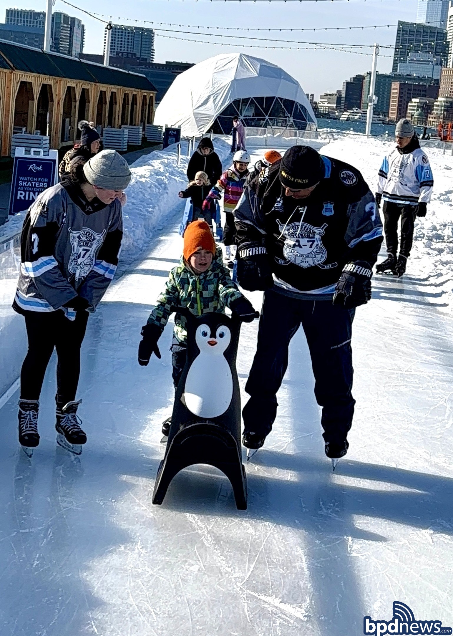 Boston Police Officer and Boston Police Cadet assist a young community member in trying to learn to skate.
